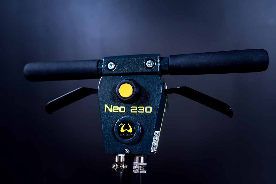 WOLFF_Application Image_Neo230_Detail3_2022-11 Close-up of the handlebar and control module of a 'Neo 230' electric scooter. The textured dark-blue metal panel features a prominent yellow circular button above a round WOLFF logo, foam grips on both sides, two black brake levers, and two heavy cables with metal connectors at the bottom. A small QR-code sticker is visible on the side. The scene is studio-lit against a dark blue-black background with sharp focus on the front control area.