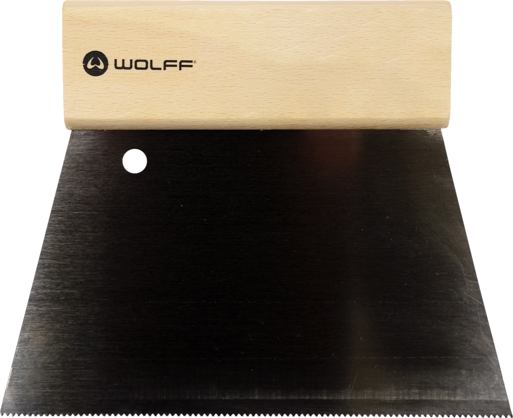 Close-up of a wide putty knife or scraper: a light rectangular wooden handle stamped with the black 'WOLFF' logo attached to a large black metal blade. The blade features a round hanging hole near the top and a fine serrated edge along the bottom; the metal shows a brushed texture. Photo taken from above on a white background.