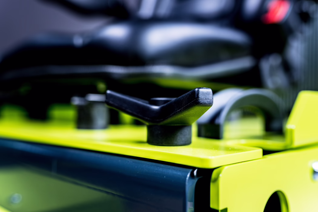 WOLFF_Application Image_Independence_2_2023-04 Close-up of a black plastic wing nut or adjustment lever mounted on a bright lime-green metal plate, part of a larger mechanical or workshop device. The shallow depth of field keeps the lever sharply in focus while the background — including a padded black component and other metal parts — is softly blurred. The image emphasizes texture, contrast and industrial detail, with visible wear and small scratches on the plastic and painted surfaces.