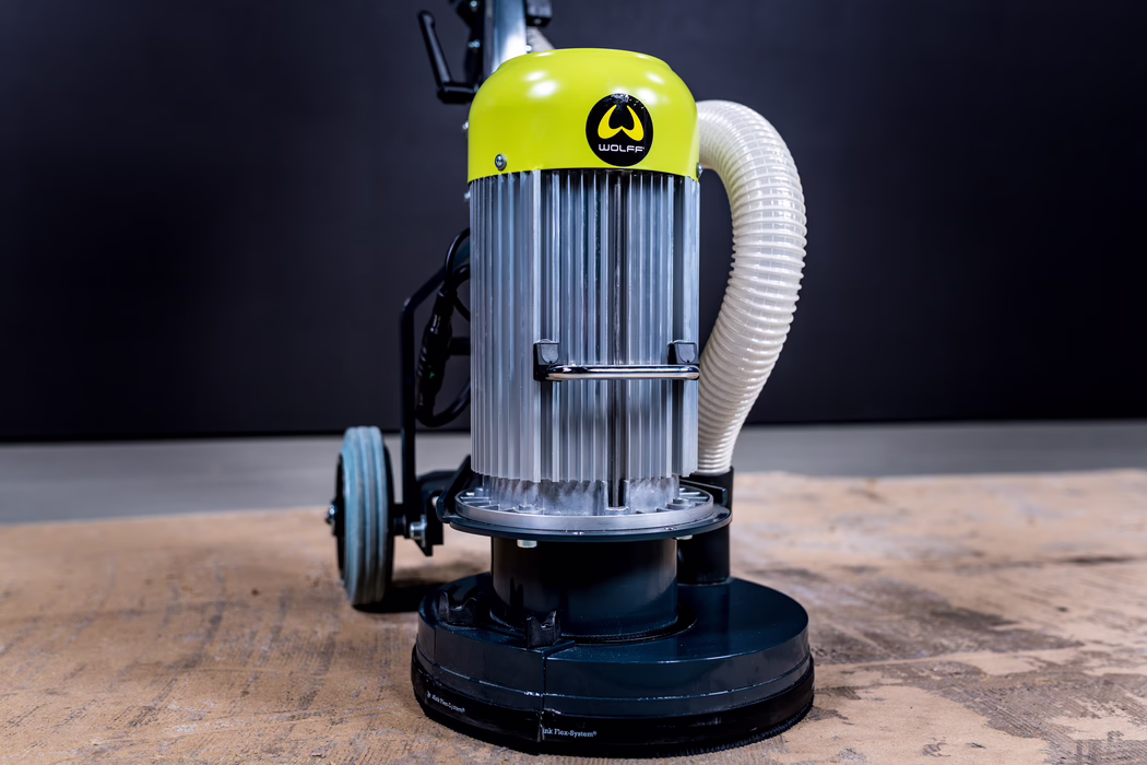 WOLFF_Application Image_Neo230_Detail7_2022-11 Close-up of a professional floor grinding and polishing machine positioned on a workshop floor. The device features a cylindrical metal motor housing with vertical cooling fins and a bright lime-green top cover displaying a 'WOLFF' logo. A flexible corrugated dust-extraction hose is attached to the side and curves upward, while the round black base houses the rotating grinding/polishing head. Two small wheels and part of the frame are visible in the background; cool, directional lighting highlights metallic textures, scratches and signs of industrial use, suggesting surface preparation or renovation work.