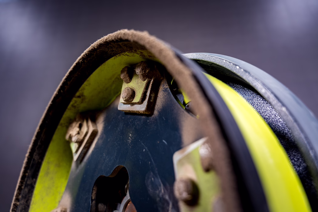 WOLFF_Application Image_Mambo_2024-01 (35) A tight close-up of a worn mechanical pulley or cable reel showing a curved metal drum with chipped neon-yellow paint, a grit-covered fabric or felt friction lining, and several rusted bolts and brackets. Soft shallow depth of field keeps the foreground edge sharp while the background fades into a smooth dark bokeh; a textured black rubber rim and a coiled yellow cable are partially visible at the right. The image emphasizes material contrasts—rough dust and rust against painted metal and rubber—and conveys an aged, industrial character.