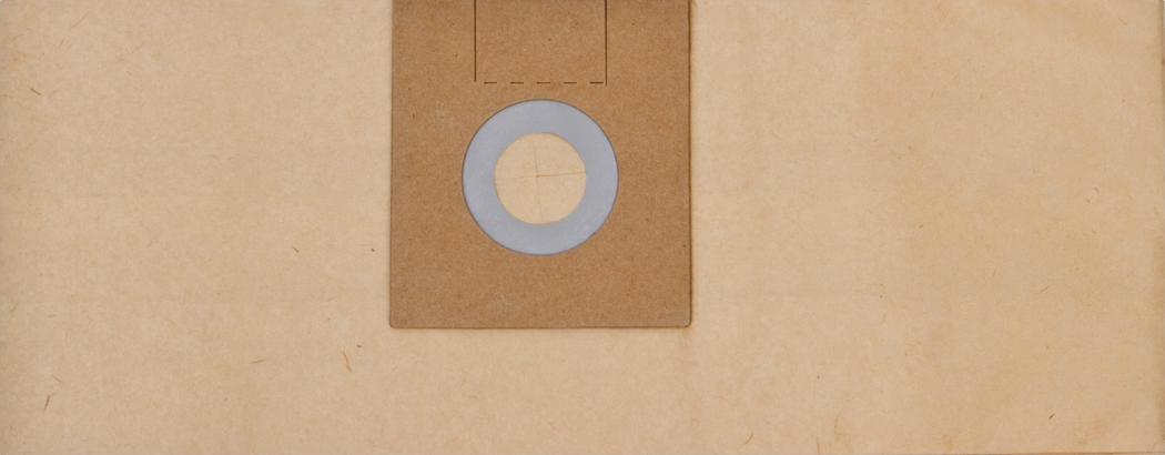 Close-up van een centraal geplaatste vierkante bruine kartonnen kraag van een stofzuigerzak op een lichtbeige, getextureerde papieren achtergrond. Het karton heeft een grijze rubberen of schuimrubberen afdichting rond een ronde opening met vier naar binnen gesneden flapjes. Het oppervlak vertoont fijne papiervezels, zacht en gelijkmatig licht en gedempte warme tinten, wat een minimalistische en tastbare compositie oplevert met lichte gebruikssporen langs de randen.