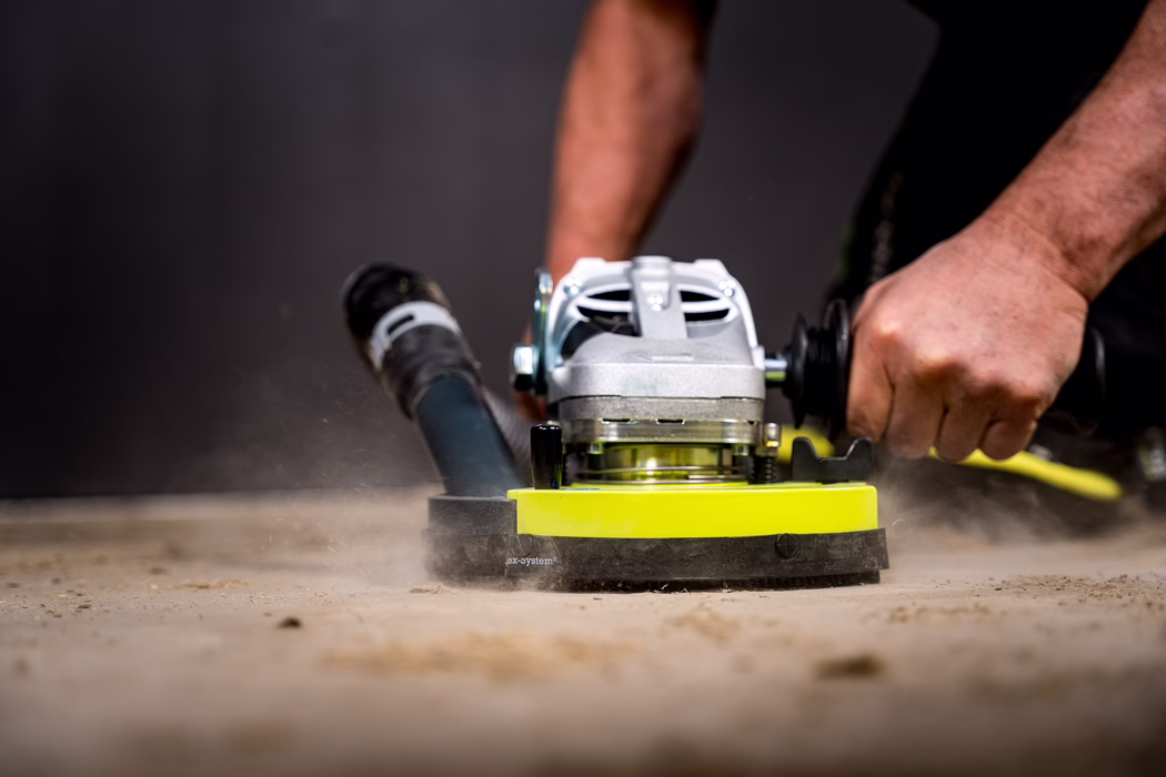 WOLFF_Application Images_BS 180_2024-01 (18) Close-up van een vakman die een draagbare vloerschuurmachine over een betonoppervlak bedient; de gele schuurkop draait en doet stof opwaaien terwijl stevige handen de handgrepen vasthouden.