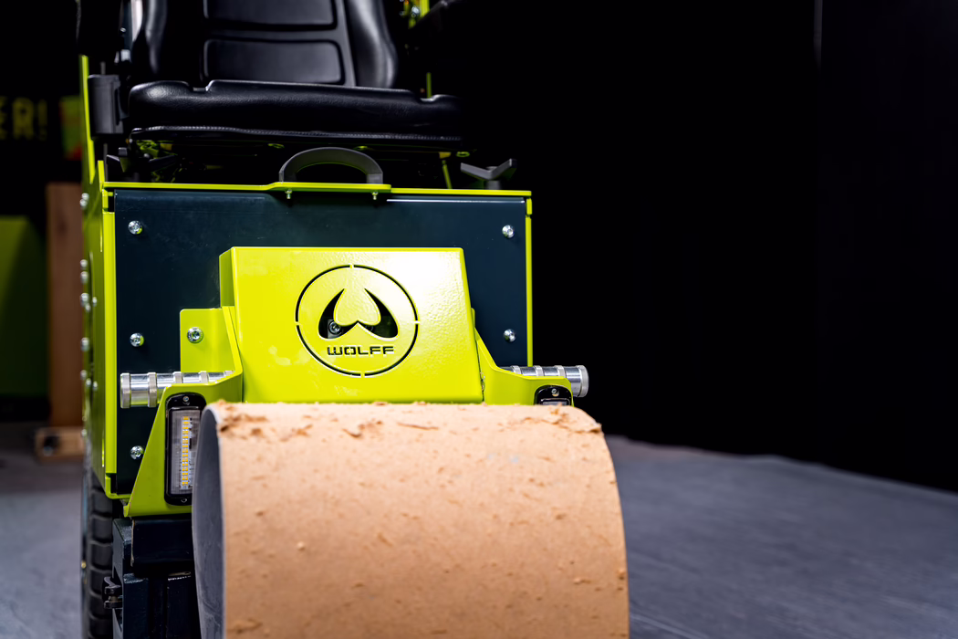 WOLFF_Application Images_Maverick_2024-01 (14) Close-up, low-angle view of the front section of a compact road roller painted in bright lime green. The image focuses on a glossy metal housing with a circular 'WOLFF' logo stamped into the panel, visible mounting screws and small LED work lights on either side. In the foreground a beige cylindrical roller with a slightly rough, textured surface occupies the lower part of the frame, while behind the housing a black padded operator seat and parts of the chassis are partially visible. The background at right is deep black, isolating the machine and emphasizing the contrast between the vivid green paint, the neutral roller material and the industrial metal details; lighting highlights surface reflections, bolts and texture, creating a sharp, technical presentation of the equipment.
