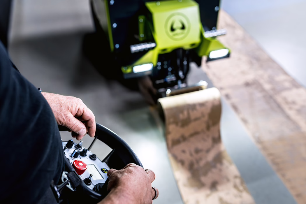 WOLFF_Application Image_Independence_14_2023-04 A close-up view of a worker operating a handheld remote control for a compact green construction machine. The image focuses on the worker's hands gripping a circular controller with several buttons, switches and a prominent red emergency-stop button, while the machine's front attachment and illuminated lights appear blurred in the background. The scene suggests a workshop or indoor construction environment with shallow depth of field, worn metal textures on the bucket and scuffed surfaces, and realistic skin details on the operator's hands. The composition emphasizes manual control, machine operation and industrial detail.