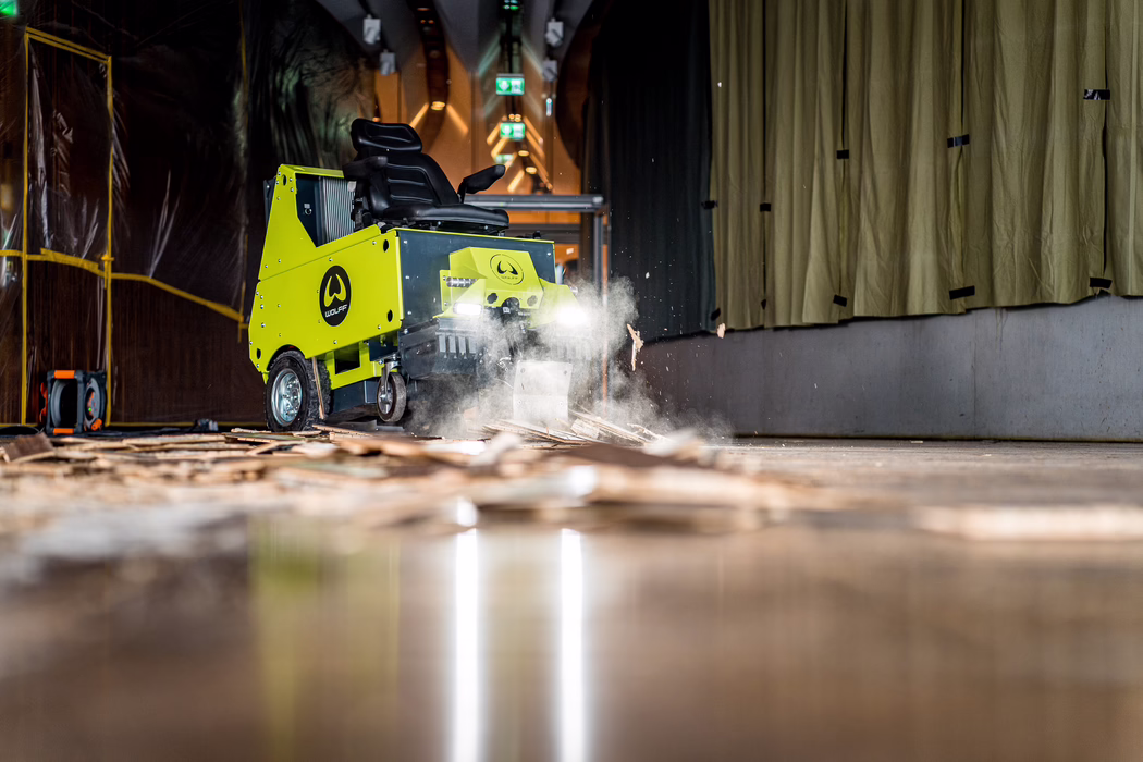 WOLFF_Application Image_Mercedes Benz Museum_2023-10 (32) Low-angle, detailed view of a bright lime-green industrial floor-scraping machine working in a dim indoor corridor. The ride-on machine, branded with a circular 'WOLFF' logo on its side, has its front blade engaged and headlights on, kicking up a cloud of dust and wood splinters as it strips old wooden flooring. Debris and broken floorboards lie scattered across the polished surface in the foreground, with sparks of material and airborne particles frozen in midair by the camera. On the left side plastic sheeting and temporary scaffolding can be seen; on the right heavy beige curtains hang from the wall. Overhead exit signs and industrial lighting create a sense of depth down the corridor. The scene emphasizes motion, texture and contrast between the vivid machine and the muted, work-in-progress environment; no people are visible and the machine appears to be in active operation.